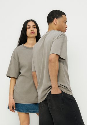 To unge voksne iført oversized grå t-shirts, den ene vendt mod kameraet i en denimnederdel, den anden vendt til siden med hænderne i lommerne på sorte jeans.