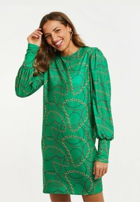 Vihreä mekko, jossa on ketjumainen kuvio, pitkät puff-hihat, korkea pääntie ja suora rakenne. Kangas on sileä ja siinä on kevyt kiilto.
