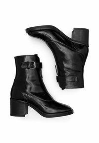 Botas de tobillo de cuero negro con un acabado brillante, tacón cuadrado y detalle de correa con hebilla. Presentan una punta redonda y una textura suave.