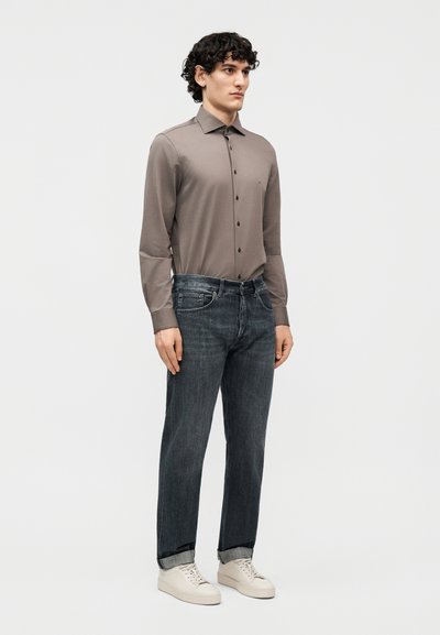 Άνδρας στέκεται φορώντας μια μακριά μπλούζα με κουμπιά σε χρώμα ταουπ, σκούρα γκρι τζιν με μανσέτες και λευκά αθλητικά παπούτσια σε απλό φόντο.