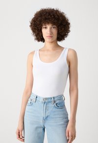 White sleeveless bodysuit with a scoop neckline, paired with light blue high-waisted jeans. Smooth fabric texture, minimal design.