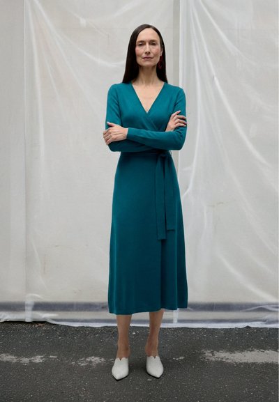 Woman with long dark hair wearing a teal long-sleeve wrap dress and white pointed shoes standing with arms crossed against a plain backdrop.