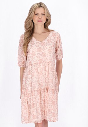 Lyshåret kvinde iført en lyserød, blomsterprintet knælængde kjole med korte ærmer og V-udskæring, stående foran en hvid baggrund.