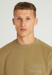 Felpa khaki con scollo rotondo e polsini a coste. Presenta testo ricamato sull'area sinistra del petto. Texture liscia e vestibilità comoda.