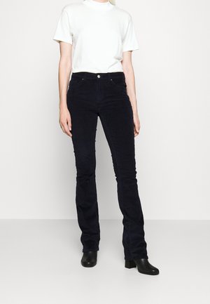 Personne portant un haut blanc à manches courtes, un pantalon en velours côtelé noir taille haute et des bottines à talons noires, debout devant un fond uni.