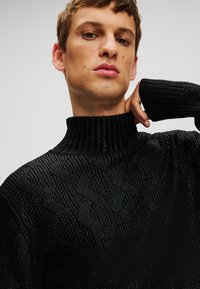 Sort strikket sweater med en høj ribbet krave og et tekstureret zigzag-mønster. Materialet har en subtil glans hele vejen igennem.