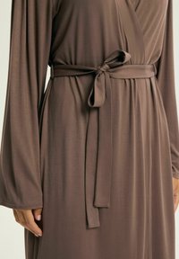 Brązowa sukienka wrap wykonana z miękkiego materiału, z długimi rękawami, paskiem wiązanym w talii oraz gładką, swobodnie opadającą tkaniną.