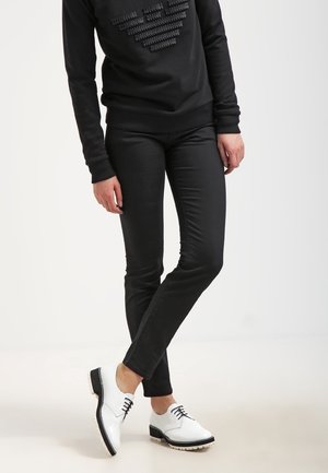 Personne portant un pantalon noir slim, un haut noir à manches longues avec motif en relief, et des chaussures blanches à lacets avec semelles noires, debout jambes croisées.