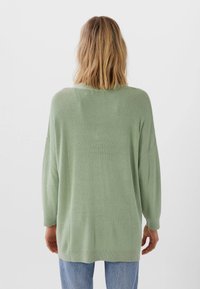 Maglione oversize verde chiaro con texture a costine, spalle cadenti e ampio scollo, abbinato a jeans blu larghi.