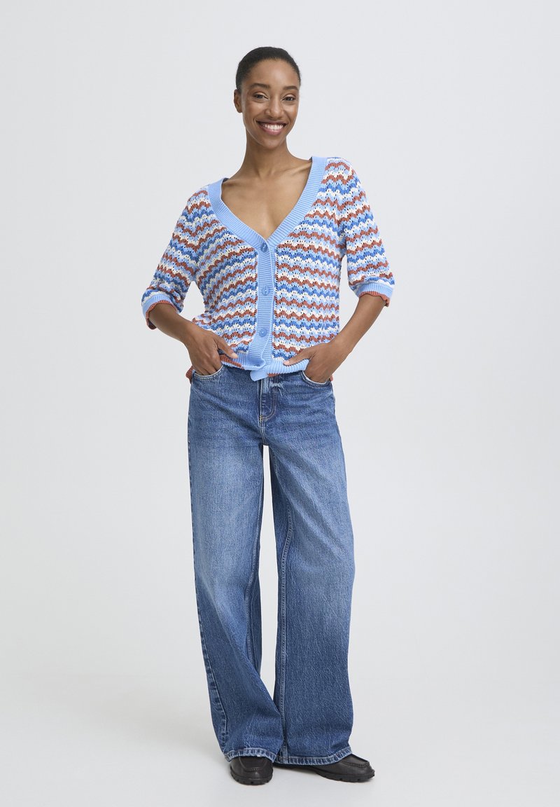 Vrouw die glimlacht, draagt een blauw-wit-oranje gestreepte cardigan, blauwe jeans met wijde pijpen en zwarte schoenen, staat met handen in de zakken.