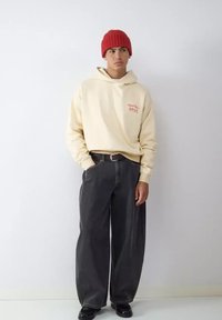 Sudadera con capucha color crema con texto rojo, combinada con jeans de corte holgado en gris oscuro. Complementada con un gorro de punto rojo y zapatos negros.