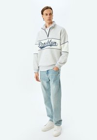 Sudadera de medio cierre en gris claro con estampado de "Brooklyn", rayas en azul marino, combinada con jeans azul claros y zapatillas blancas.
