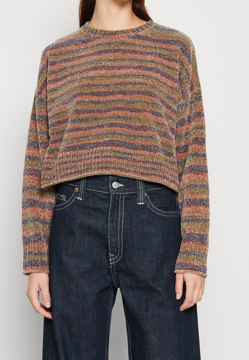 Pull en maille rayé aux tons terre atténués, associé à un jean taille haute bleu foncé avec coutures blanches visibles et fermeture par boutons.