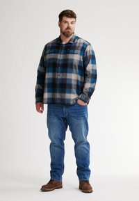 Blauw en grijs geruite flanellen overhemd met twee borstzakken, knoopsluiting aan de voorkant, gedragen met blauwe jeans en bruine werkschoenen.