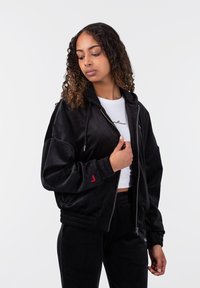 Juodas zip-up bomber tipo džemperis su minkšta tekstūra, turintis gipiūrines apykakles, gobtuvą ir mažą raudoną logotipą kairiajame rankovėje.