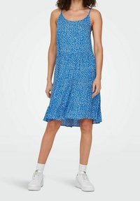 Sininen kukallinen mekko ohuilla olkaimilla, polvipituinen, valuvasta kankaasta, jossa on hajanaisia valkoisia kukkakuviota, yhdistetty valkoisiin tennareihin.