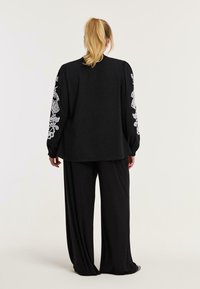 Schwarzes Langarmshirt mit weißen, gestickten Details an den Ärmeln, kombiniert mit locker sitzenden schwarzen Hosen. Weicher Stoff und entspannte Silhouette.