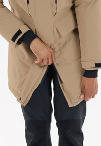 Beige vedenkestävä takki, jossa on vetoketjukiinnitys, tarranauha-yksityiskohdat ja taskut, käytetty mustien housujen päällä. Kädet tarttuvat takin etuosaan.