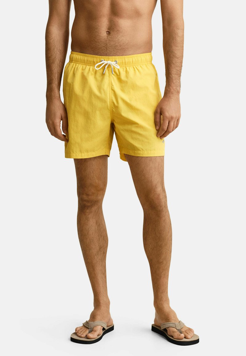 Homme portant un short de bain jaune avec un cordon blanc et des tongs beiges, debout devant un fond clair uni.