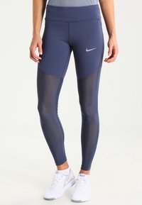 Mörkblå leggings med hög midja, med en meshpaneldesign och en silverfärgad Nike-logotyp på vänster lår. Mjukt, elastiskt material.