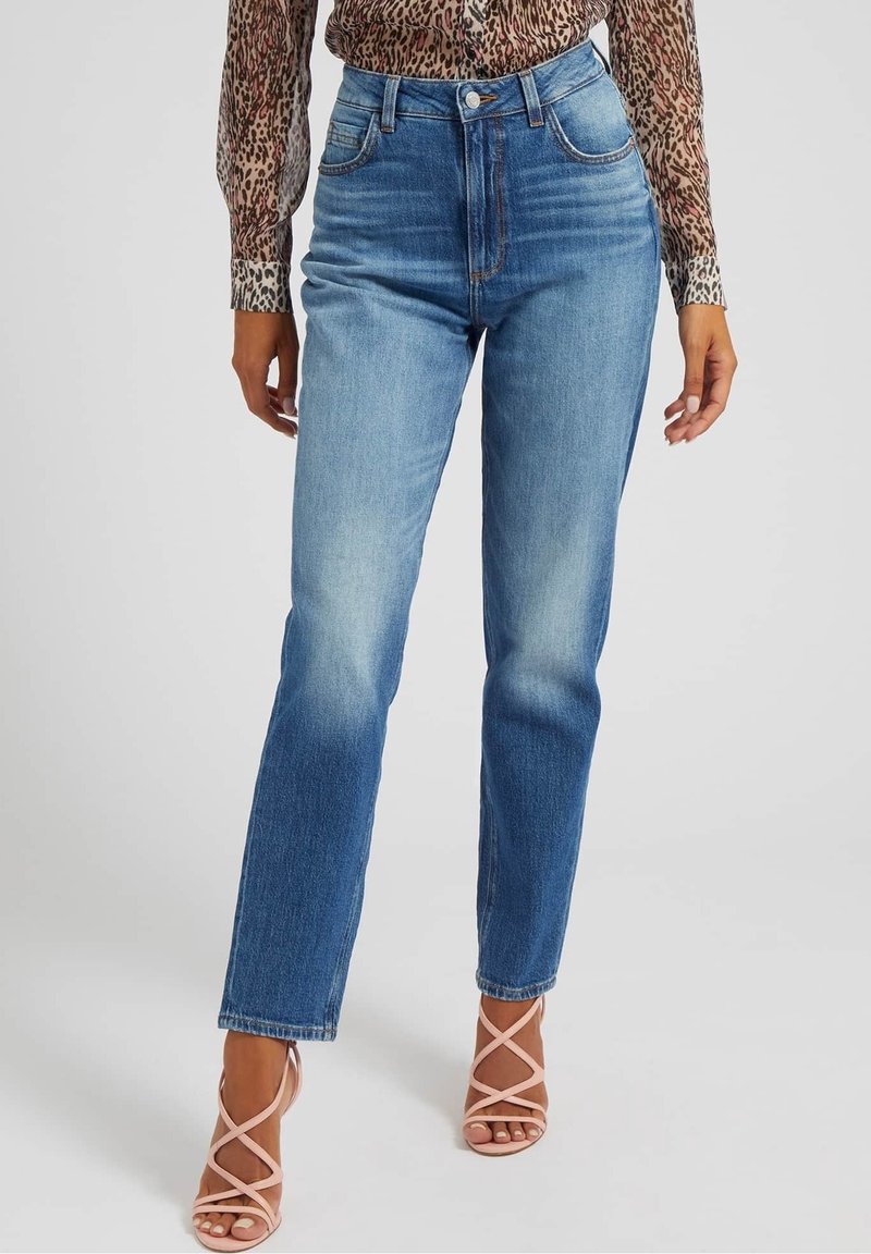 Femme portant un jean bleu taille haute avec des détails délavés, associé à un chemisier à manches longues imprimé léopard et des sandales à talons rose à lanières.