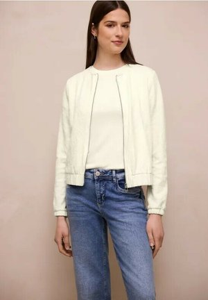 Femme portant une veste blanche texturée zippée sur une chemise blanche, associée à un jean bleu taille haute, debout devant un fond uni.