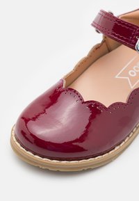 Bordeauxfarbene Lackleder-Schuhe mit gewelltem Rand, verstellbarem Riemen und einer beigefarbenen Gummisohle, die eine glatte und glänzende Oberfläche aufweisen.