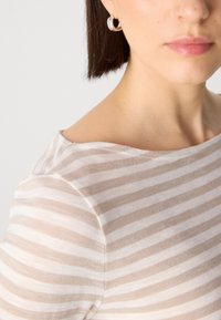 Stripetopp i lys beige og hvit, laget av et mykt, gjennomsiktig stoff med off-shoulder halslinje og synlige sømdetaljer. Sølvfargede ringøredobber.