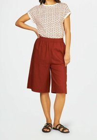 Femme portant un short large de couleur rouille, un haut à manches courtes à motif et des sandales noires à lanières, se tenant les mains sur les hanches.