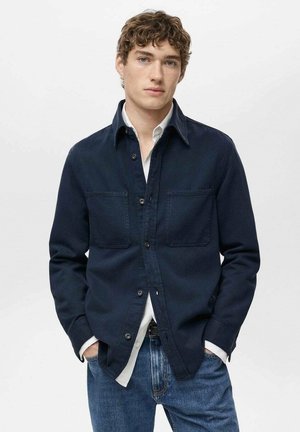 Camicia blu navy con due tasche sul petto, abbinata a una camicia bianca con colletto, tessuto strutturato e vestibilità classica, indossata con jeans blu.