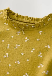 Top color senape con scollo arricciato. Presenta un motivo floreale in rosa e bianco. Accentuato da un piccolo fiocco di nastro al colletto.