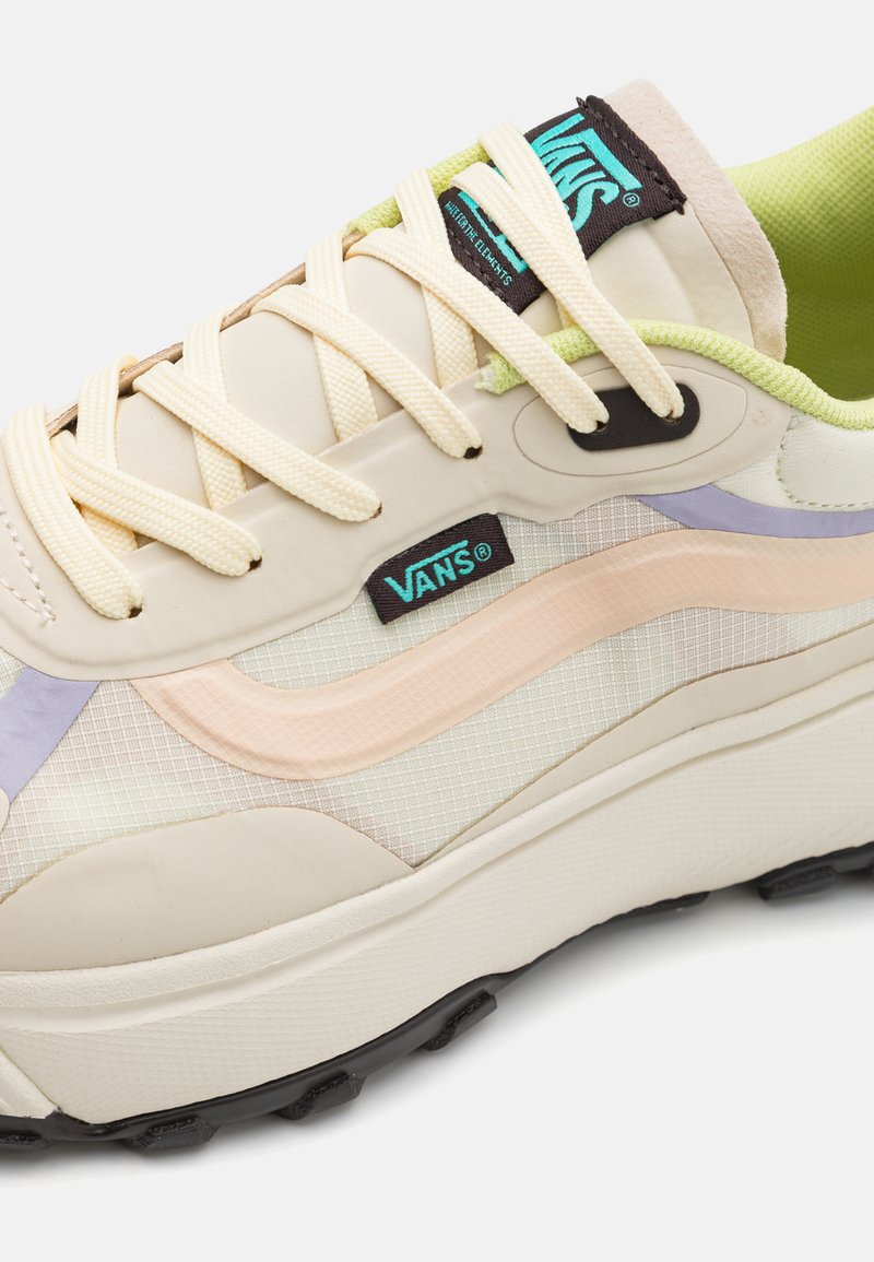 Zapatillas beige con materiales de malla y goma, con acentos rosa y morado, cordones redondos y una suela negra con caminos elevados.