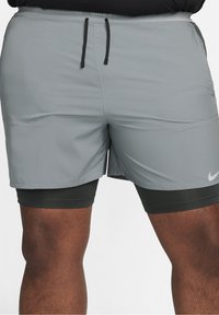 Shorts de sport gris avec cordons noirs portés par-dessus des shorts de compression noirs sur une personne debout devant un fond uni.