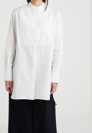 Kobieta w białej, zapinanej na guziki koszuli z długim rękawem i stójką oraz w czarnych szerokich spodniach na tle jednolitego tła.