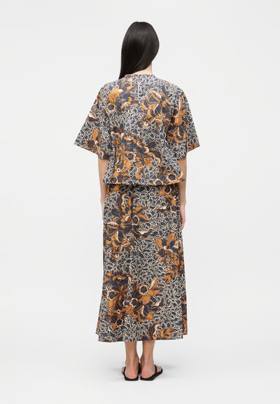 Γυναίκα με μακριά σκούρα μαλλιά, φοράει καφέ, μπλε και λευκό τοπ και μακριά φούστα με λουλουδάτο σχέδιο, στέκεται με την πλάτη στην κάμερα.