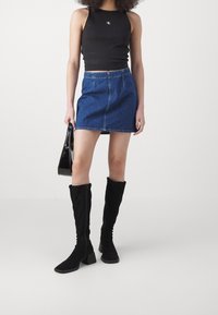 Kvinna iklädd svart ärmlös crop top, blå denim minikjol, svarta knähöga stövlar, håller en glänsande svart handväska mot vit bakgrund.