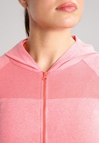 Frau in einem rosa-korallfarbenen Kapuzenpullover mit Reißverschluss, der den Ausschnitt, die Lippen und den unteren Teil des Gesichts vor einem schlichten hellen Hintergrund zeigt.