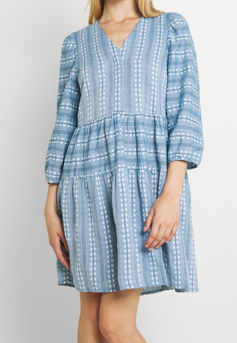Robe bleue à motifs avec un col en V, des manches ballon et une jupe à volants. Confectionnée en tissu léger avec des rayures florales blanches.