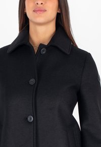 Cappotto in misto lana nera con colletto a punta, chiusura frontale a tre bottoni e dettaglio del logo sulla manica. Tessuto liscio e vestibilità sartoriale.