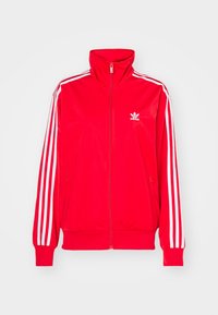 Czerwona kurtka z poliestru z wysokim kołnierzem, pełnym zamkiem z przodu, białymi trzema paskami na rękawach i małym białym logo na piersi.