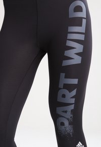 Mustat legginsit, jotka on valmistettu joustavasta kankaasta ja joissa on suuri "PART WILD" -teksti harmaana sivussa sekä pieni logodetaili nilkassa.