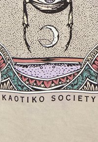 Béžové tričko s multikolorovým grafickým designem, na kterém je centrální oko, měsíc v kvádru a dekorativní vzory. Text zní "KAOTIKO SOCIETY."