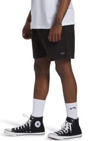 Pantalones cortos de mezclilla negros con un logo de parche, llevados con calcetines blancos hasta la altura del tobillo y zapatillas altas negras con un logo de estrella blanca.