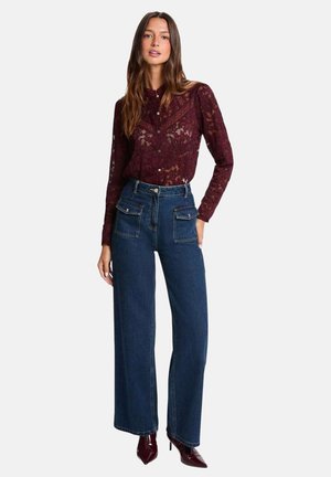 Femme portant un chemisier en dentelle bordeaux transparent à manches longues, rentré dans un jean bleu à taille haute, jambes larges, avec poches à rabat sur le devant.