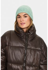 Žena má na sobě světle zelenou pletenou čepici a tmavohnědou lesklou péřovou bundu s vysokým límcem zipem zapnutým.