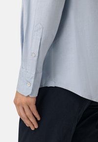 Chemise bleu clair à manches longues avec des poignets boutonnés et un ourlet pointu. Fabriquée dans un tissu lisse, avec une texture subtile. Pantalons foncés visibles en dessous.