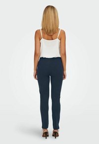 Marinblå slim-fit byxor i stretchtyg med hög midja och ankellång design. Kombinerade med en vit topp med spaghetti-band och klackar.