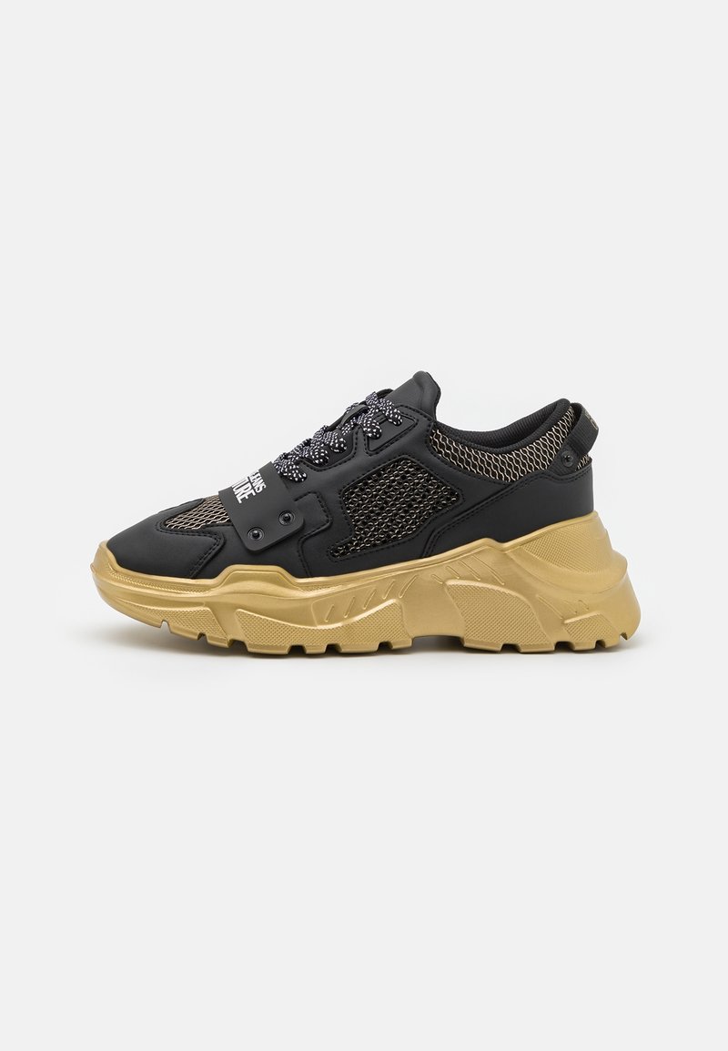 Zapatilla gruesa negra y dorada con paneles de malla, cordones estampados y una suela gruesa y texturizada, mostrada de lado sobre un fondo blanco.