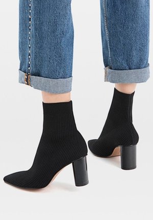 Bottes chaussettes noires côtelées avec talons carrés portées avec des jeans bleus retroussés, montrant un détail de couture latérale cloutée.