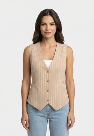 Vrouw met lang bruin haar, die een beige mouwloos vest met krijtstreep draagt over een wit shirt, gecombineerd met lichtblauwe jeans, staand tegen een witte achtergrond.